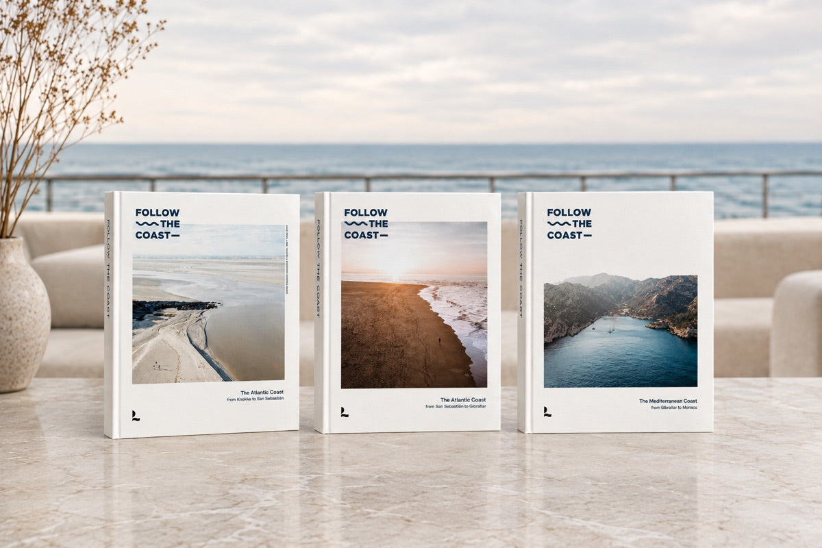 Three white books titled 'FOLLOW THE COAST' are standing upright on a light stone surface in a bright, modern interior with a blurred ocean and sofa background.